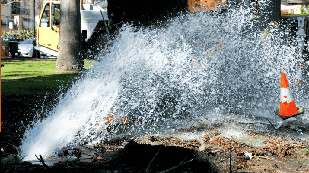 Water bursts forcefully from a broken underground pipe in a park, spraying high into the air. A traffic cone stands nearby, and a utility truck is visible in the background.
