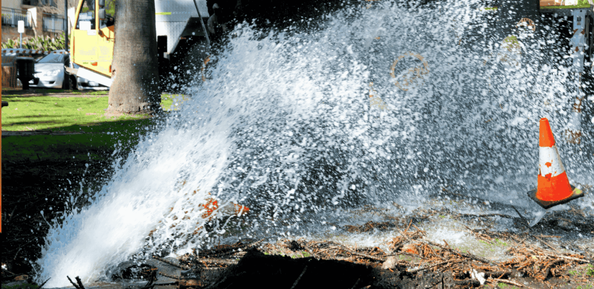 Water bursts forcefully from a broken underground pipe in a park, spraying high into the air. A traffic cone stands nearby, and a utility truck is visible in the background.