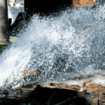Water bursts forcefully from a broken underground pipe in a park, spraying high into the air. A traffic cone stands nearby, and a utility truck is visible in the background.