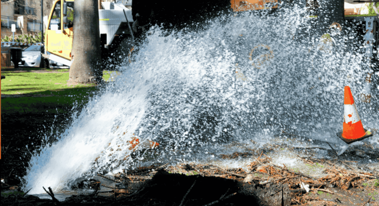 Water bursts forcefully from a broken underground pipe in a park, spraying high into the air. A traffic cone stands nearby, and a utility truck is visible in the background.