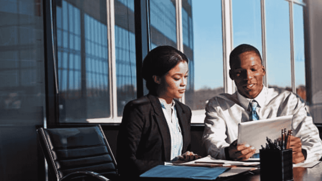 Two business professionals sit at a desk in a modern office with large windows, reviewing strategies on a digital tablet to address potential infrastructure collapse. Papers and office supplies are spread out on the table.