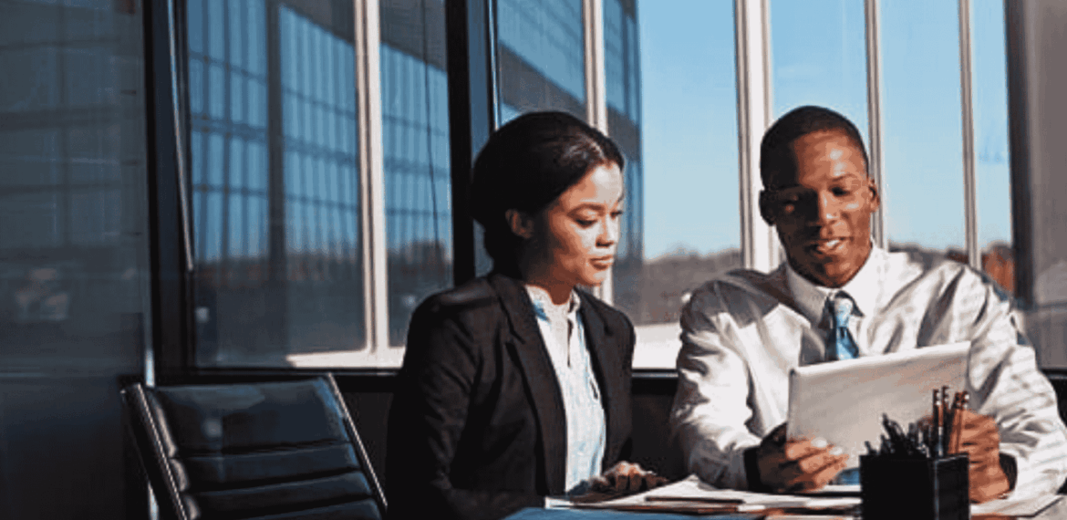 Two business professionals sit at a desk in a modern office with large windows, reviewing strategies on a digital tablet to address potential infrastructure collapse. Papers and office supplies are spread out on the table.