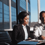 Two business professionals sit at a desk in a modern office with large windows, reviewing strategies on a digital tablet to address potential infrastructure collapse. Papers and office supplies are spread out on the table.