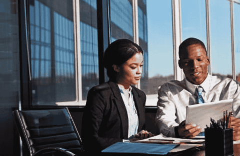 Two business professionals sit at a desk in a modern office with large windows, reviewing strategies on a digital tablet to address potential infrastructure collapse. Papers and office supplies are spread out on the table.