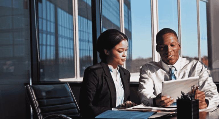 Two business professionals sit at a desk in a modern office with large windows, reviewing strategies on a digital tablet to address potential infrastructure collapse. Papers and office supplies are spread out on the table.