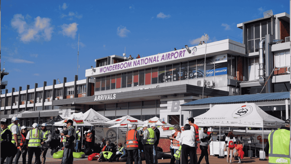 A busy scene outside Wonderboom National Airport shows people in safety vests, tents, and booths under a sunny sky. The airport building has a sign reading WONDERBOOM NATIONAL AIRPORT above the arrivals area.