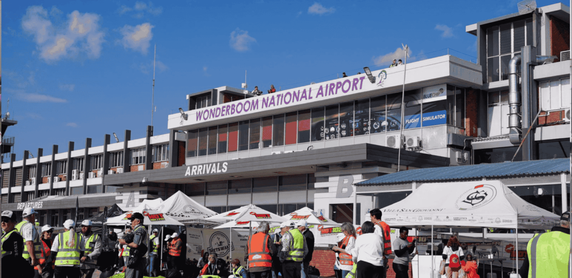 A busy scene outside Wonderboom National Airport shows people in safety vests, tents, and booths under a sunny sky. The airport building has a sign reading WONDERBOOM NATIONAL AIRPORT above the arrivals area.