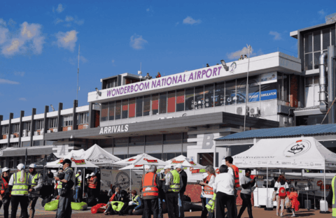 A busy scene outside Wonderboom National Airport shows people in safety vests, tents, and booths under a sunny sky. The airport building has a sign reading WONDERBOOM NATIONAL AIRPORT above the arrivals area.