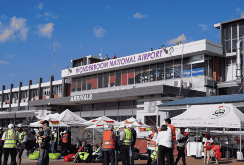 A busy scene outside Wonderboom National Airport shows people in safety vests, tents, and booths under a sunny sky. The airport building has a sign reading WONDERBOOM NATIONAL AIRPORT above the arrivals area.