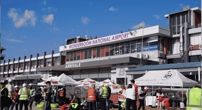 A busy scene outside Wonderboom National Airport shows people in safety vests, tents, and booths under a sunny sky. The airport building has a sign reading WONDERBOOM NATIONAL AIRPORT above the arrivals area.