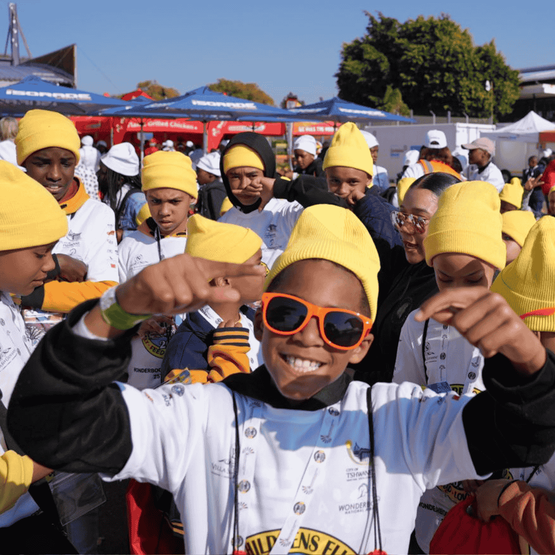 A group of smiling children wearing yellow beanies and white shirts gather outdoors at an event. One child in the front wears orange sunglasses and points happily at the camera. People and tents are visible in the background.