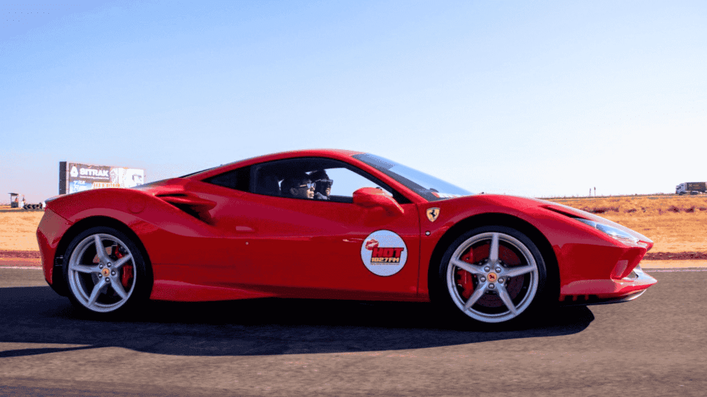 A bright red Ferrari sports car with a Hot Lap sticker on the door drives on a racetrack with dry grass and a clear blue sky in the background.