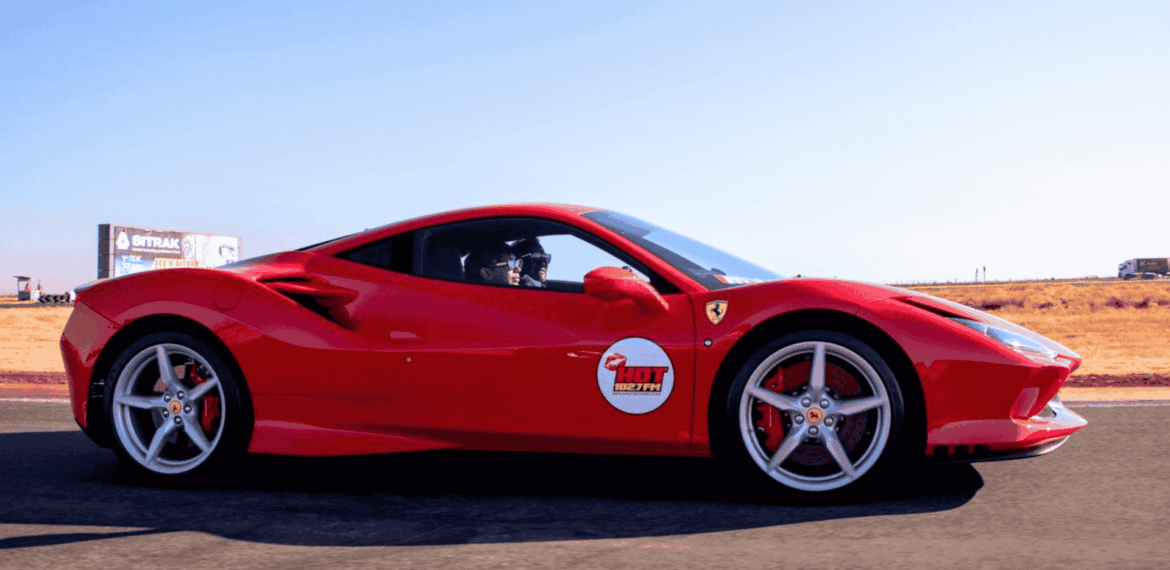 A bright red Ferrari sports car with a Hot Lap sticker on the door drives on a racetrack with dry grass and a clear blue sky in the background.