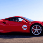 A bright red Ferrari sports car with a Hot Lap sticker on the door drives on a racetrack with dry grass and a clear blue sky in the background.