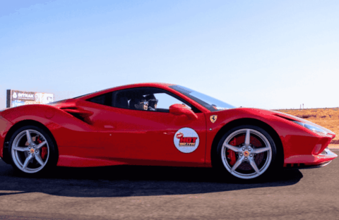 A bright red Ferrari sports car with a Hot Lap sticker on the door drives on a racetrack with dry grass and a clear blue sky in the background.