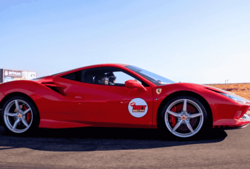 A bright red Ferrari sports car with a Hot Lap sticker on the door drives on a racetrack with dry grass and a clear blue sky in the background.