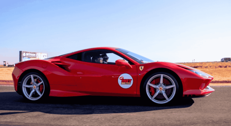 A bright red Ferrari sports car with a Hot Lap sticker on the door drives on a racetrack with dry grass and a clear blue sky in the background.