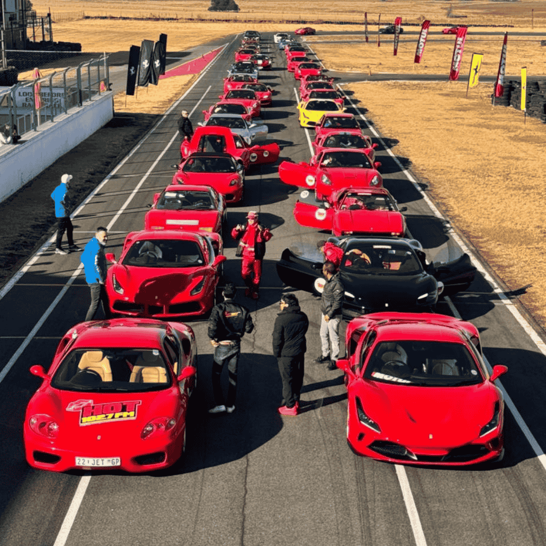 A lineup of red sports cars, mostly Ferraris, parked closely on a racetrack. People stand and walk between the cars, some with open doors, on a sunny day with dry grass in the background.
