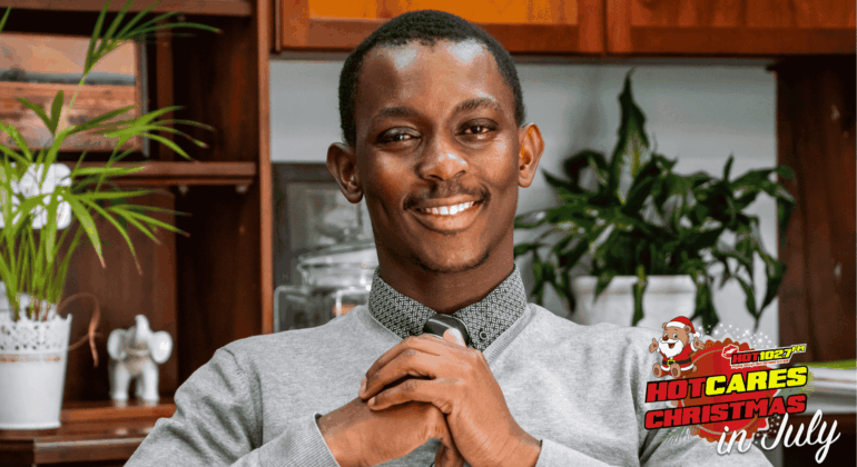 A smiling man in a grey sweater and tie sits with hands clasped. Behind him are plants and wooden shelves. The image has a Hot Cares Christmas in July logo in the bottom right corner.