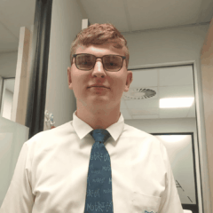 A young man with short light brown hair and glasses wears a white shirt and a blue tie with text. He is standing indoors in a brightly lit office environment.