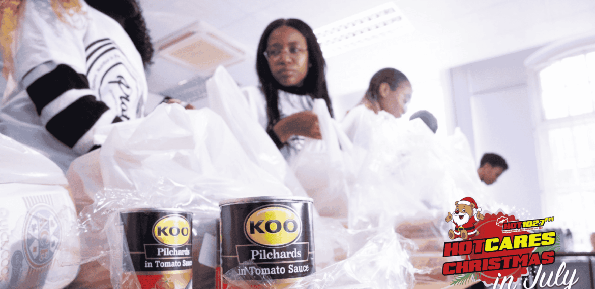 People packing food items, including KOO pilchards cans, into plastic bags on a table. The image features a HOT CARES CHRISTMAS in July logo with a Santa illustration in the bottom right corner.