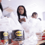 People packing food items, including KOO pilchards cans, into plastic bags on a table. The image features a HOT CARES CHRISTMAS in July logo with a Santa illustration in the bottom right corner.