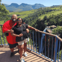 A smiling couple stands together on a viewpoint balcony overlooking a lush green landscape and a tall waterfall, with mountains visible in the background under a clear blue sky.