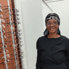 A woman wearing glasses and a black headscarf stands indoors next to a display rack filled with various eyeglasses. She is smiling and dressed in black.