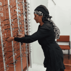 A woman in a black dress and patterned headscarf selects eyeglasses from a display rack in a store. There is a brown chair and white walls in the background.