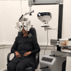 A person sits in an eye examination chair wearing a phoropter while undergoing a vision test in an optometrist’s office. Ophthalmic equipment and a control panel are visible beside them.