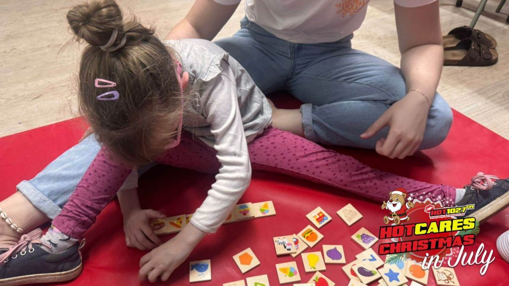 A young girl sits on a red mat playing with colorful wooden tiles while an adult sits beside her. The image has a HOT 102.7 FM Hot Cares Christmas in July logo in the bottom right corner.