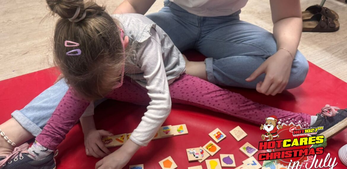 A young girl sits on a red mat playing with colorful wooden tiles while an adult sits beside her. The image has a HOT 102.7 FM Hot Cares Christmas in July logo in the bottom right corner.