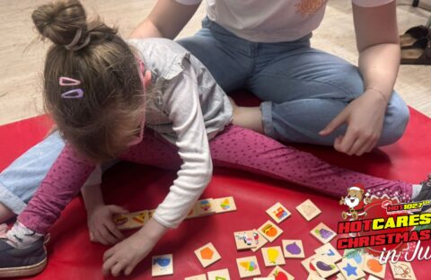A young girl sits on a red mat playing with colorful wooden tiles while an adult sits beside her. The image has a HOT 102.7 FM Hot Cares Christmas in July logo in the bottom right corner.
