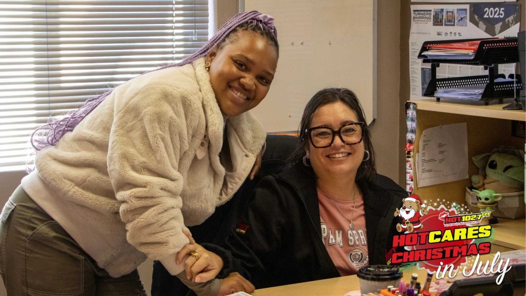 Two women smiling in an office, one standing and one sitting at a desk. A festive HOT CARES Christmas in July logo is displayed in the bottom right corner.