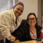 Two women smiling in an office, one standing and one sitting at a desk. A festive HOT CARES Christmas in July logo is displayed in the bottom right corner.