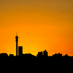 Silhouette of a city skyline with a tall tower at sunset, featuring an orange sky that gradually fades into yellow near the horizon.