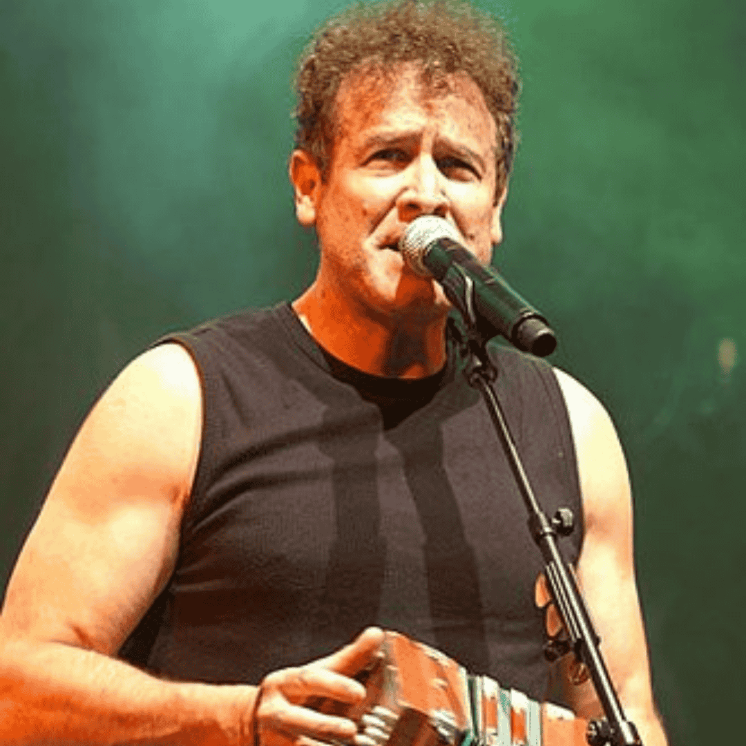 A man with curly hair and a sleeveless black shirt sings into a microphone on stage, holding an accordion, with green stage lighting in the background, evoking the raw energy of classic mixtapes.