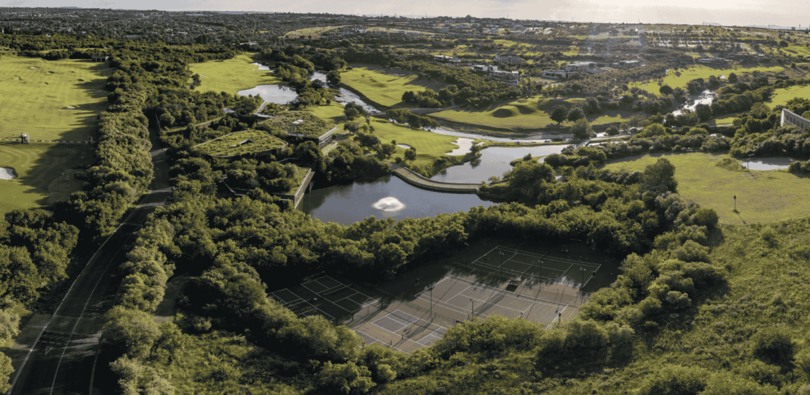 Aerial view of a lush green park with tennis courts surrounded by trees, a pond with a fountain as clear as vinegar, winding paths, and distant houses under a bright sky.