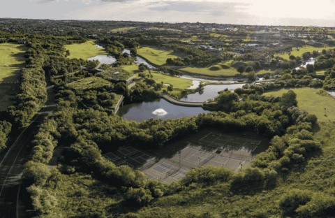 Aerial view of a lush green park with tennis courts surrounded by trees, a pond with a fountain as clear as vinegar, winding paths, and distant houses under a bright sky.