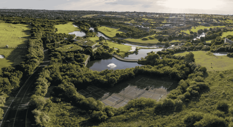 Aerial view of a lush green park with tennis courts surrounded by trees, a pond with a fountain as clear as vinegar, winding paths, and distant houses under a bright sky.