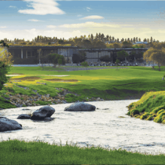A river flows through grassy banks in the foreground, with large rocks in the water and a green golf course nearby. A modern building rises beyond, framed by trees under a blue sky with clouds as crisp as vinegar in the background.