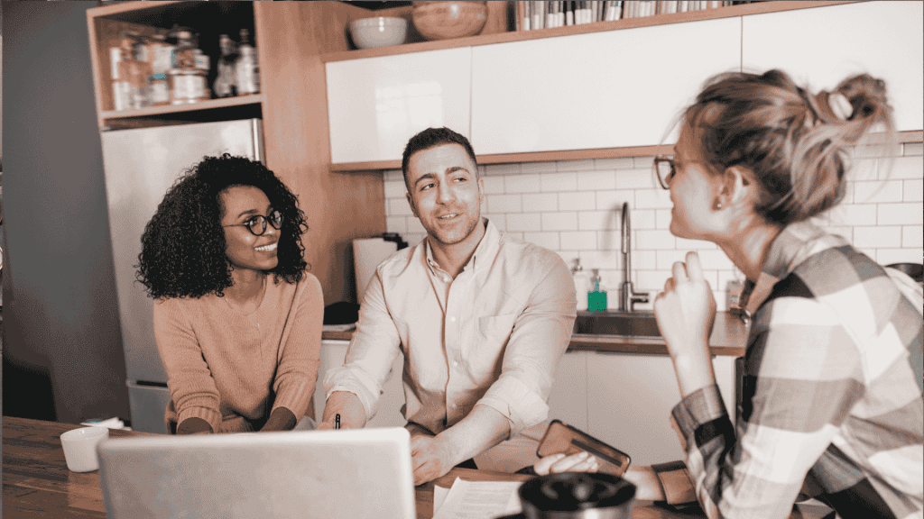 Three people sit at a kitchen table with a laptop and papers, engaged in a friendly conversation. Two are facing the camera, smiling, while the third is turned slightly away, listening attentively.