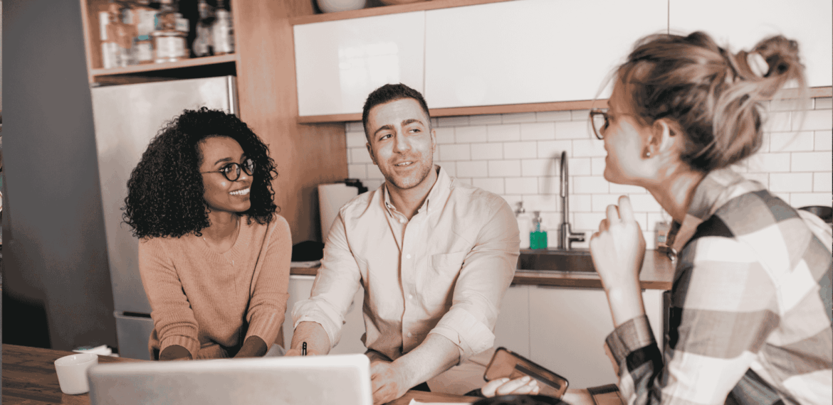 Three people sit at a kitchen table with a laptop and papers, engaged in a friendly conversation. Two are facing the camera, smiling, while the third is turned slightly away, listening attentively.