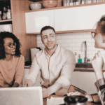 Three people sit at a kitchen table with a laptop and papers, engaged in a friendly conversation. Two are facing the camera, smiling, while the third is turned slightly away, listening attentively.