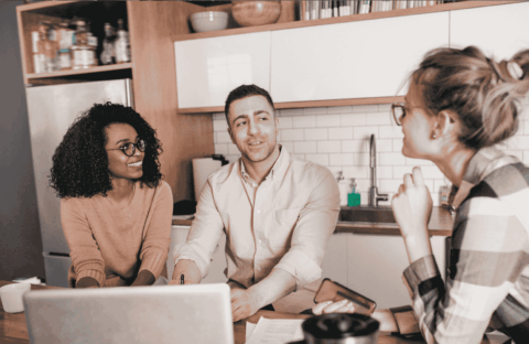 Three people sit at a kitchen table with a laptop and papers, engaged in a friendly conversation. Two are facing the camera, smiling, while the third is turned slightly away, listening attentively.