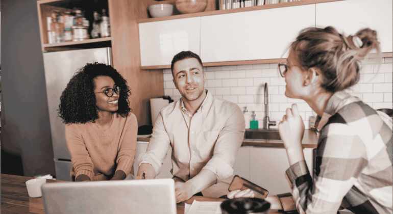 Three people sit at a kitchen table with a laptop and papers, engaged in a friendly conversation. Two are facing the camera, smiling, while the third is turned slightly away, listening attentively.