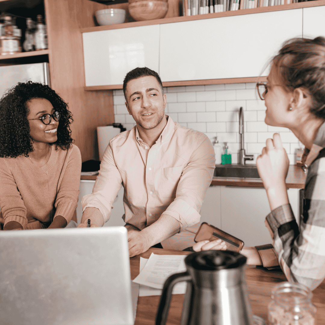 Three people sit around a kitchen table having a discussion. A laptop, documents, and a coffee pot are on the table. They appear to be collaborating or having a casual meeting in a modern kitchen setting.
