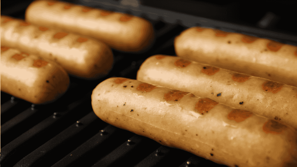 Six sausages with visible grill marks are cooking on a black grill. The sausages are light brown and evenly spaced, with a close-up focus highlighting their texture and the heat of the grill.