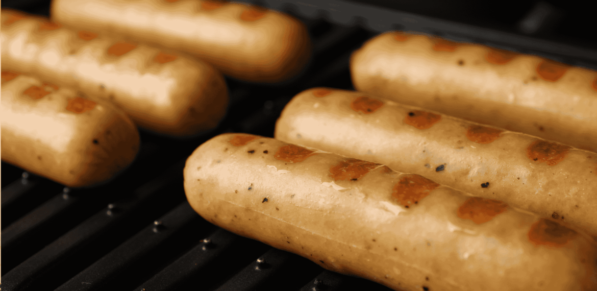 Six sausages with visible grill marks are cooking on a black grill. The sausages are light brown and evenly spaced, with a close-up focus highlighting their texture and the heat of the grill.