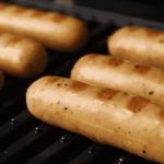 Six sausages with visible grill marks are cooking on a black grill. The sausages are light brown and evenly spaced, with a close-up focus highlighting their texture and the heat of the grill.